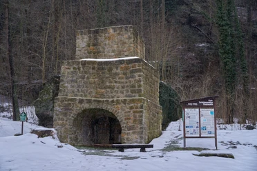 A historic blast furnace made of stone, surrounded by a snowy winter landscape with an information board in the foreground.