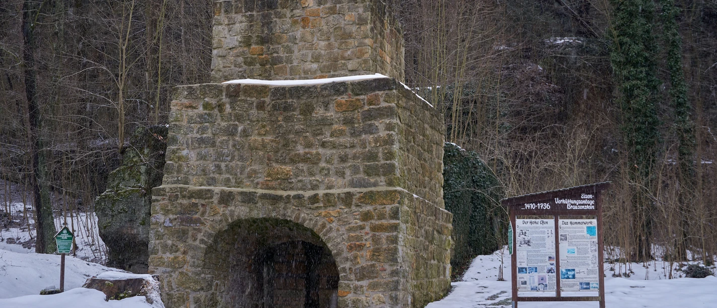 Hochofen im Winter Ein historische Hochofen aus Stein, umgeben von verschneiter Winterlandschaft mit Infotafel im Vordergrund.