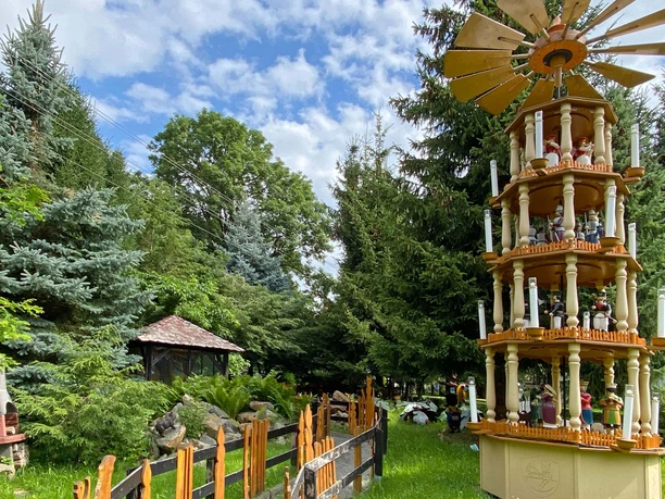 Aussenpyramide.jpg Velká několikapatrová vánoční pyramida s postavami a svíčkami v zelené zahradě s jedlemi a modrou oblohou v pozadí.
