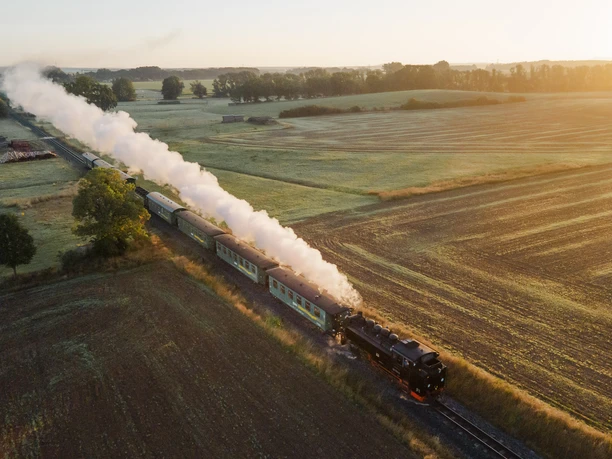 A historic steam train travels through a vast landscape in the morning light.