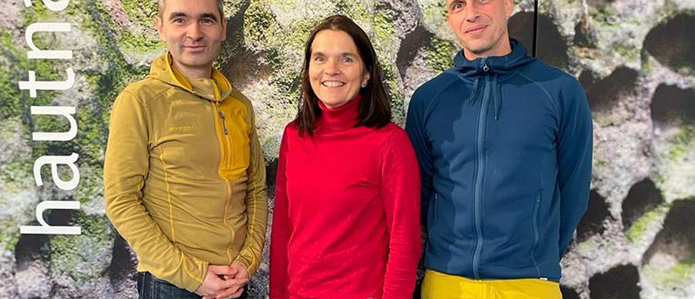 Three people stand smiling in front of a moss-covered wall. The person on the left is wearing a yellow jacket, the one in the middle a red one and the one on the right a blue one.