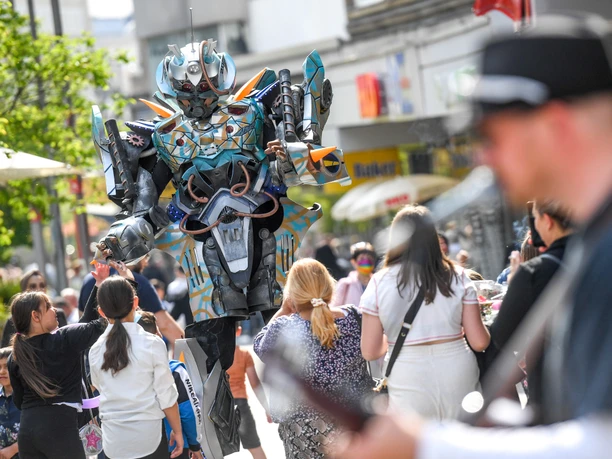 Walking Act bei Mülheim mittendrin 2022 Ein imposanter Roboter in leuchtendem Kostüm unterhält Passanten auf belebter Einkaufsstraße.