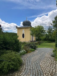 Blaues Häusel Maxen Ein gelbes Gebäude mit Kuppeldach und Halbmondsymbol, umgeben von grünen Büschen und einem geschwungenen Pflasterweg unter blauem Himmel mit Wolken.