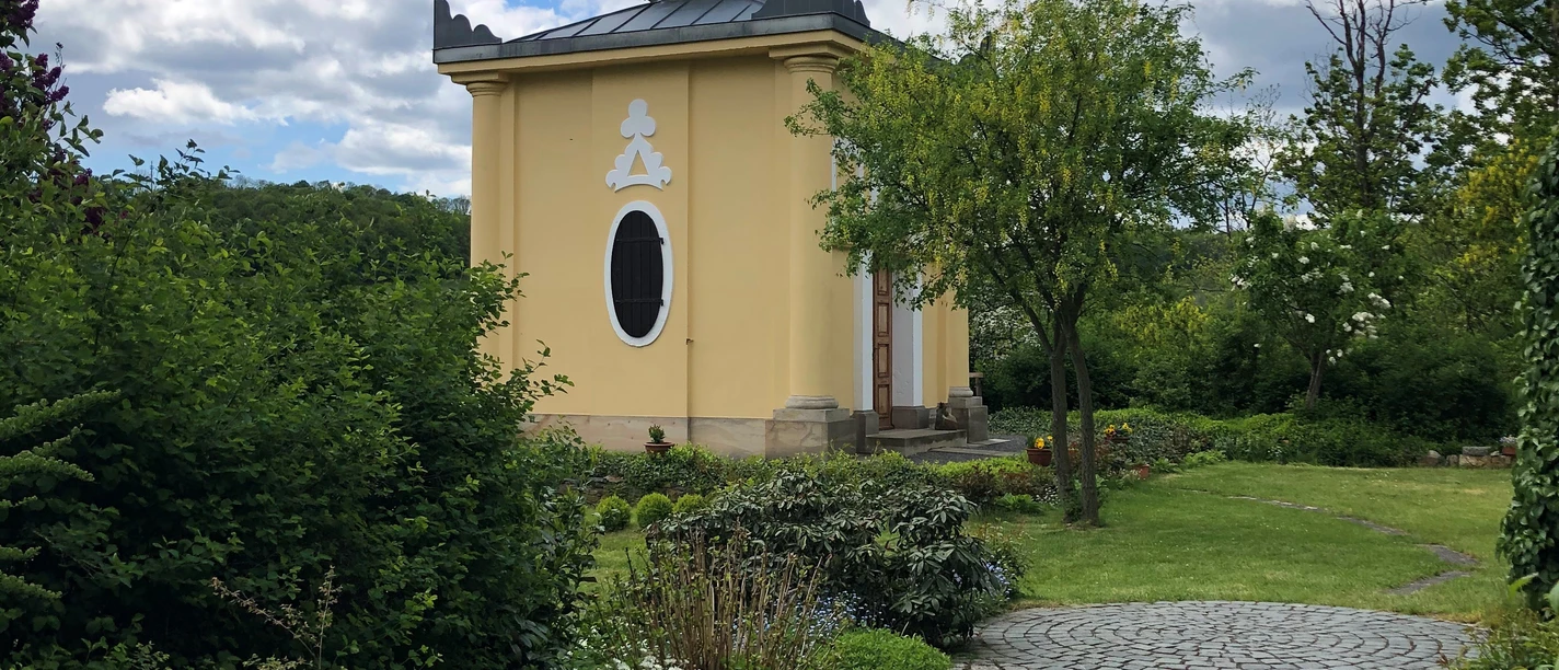 Blaues Häusel Maxen Ein gelbes Gebäude mit Kuppeldach und Halbmondsymbol, umgeben von grünen Büschen und einem geschwungenen Pflasterweg unter blauem Himmel mit Wolken.