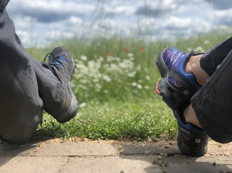 Two people in hiking gear relax on a path surrounded by green grass and blooming flowers.