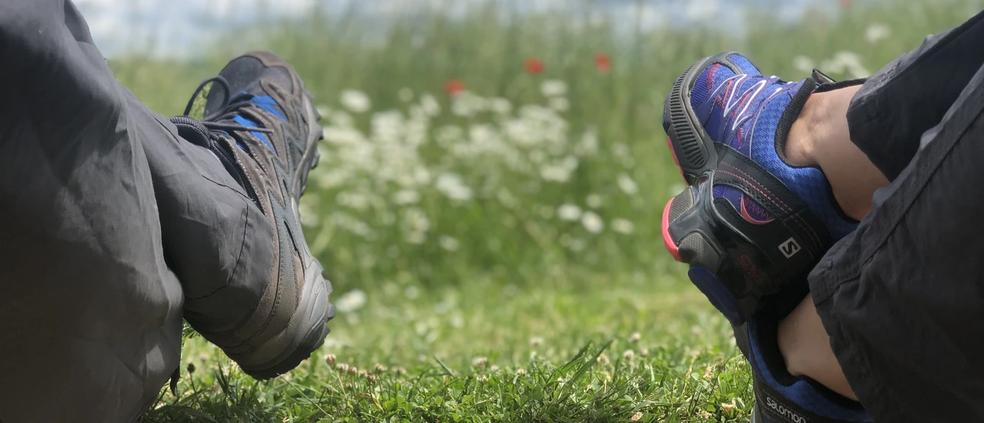 Wandern Zwei Personen in Wanderkleidung entspannen auf einem Weg, umgeben von grünem Gras und blühenden Blumen.