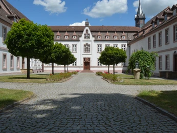 Historisches Kloster mit gepflegtem Innenhof, Bäumen und zentrale Kirche im Hintergrund.