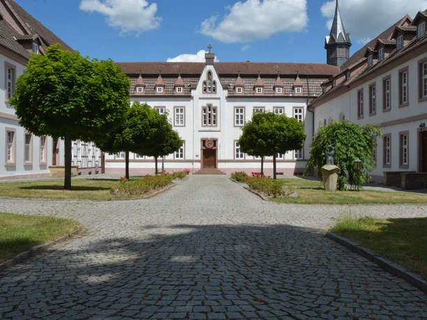 Historisches Kloster mit gepflegtem Innenhof, Bäumen und zentrale Kirche im Hintergrund.