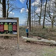 Auf der Familienvariante des Paderborner Höhenwegs Ein Kind balanciert auf einem liegenden Baumstamm neben einer Infotafel im Wald.