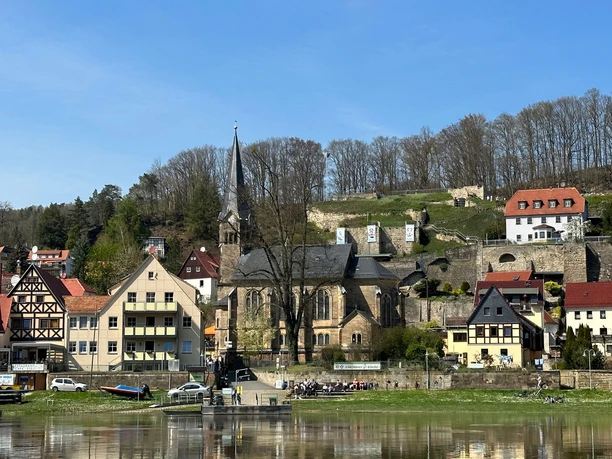 Small town view with half-timbered houses, church with high tower and river in the foreground, with a clear sky and spring-like atmosphere.
