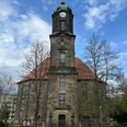 Kirche Lohmen Historische Kirche mit rotem Dach und hohem Uhrturm, umgeben von kahlen Bäumen, vor blauem Himmel mit Wolken.