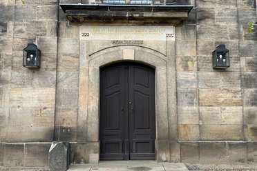 Tür der Kirche in Lohmen Dunkle Holztür in einem steinernen Gebäude, flankiert von zwei Laternen; darüber eine Inschrift in goldener Schrift.