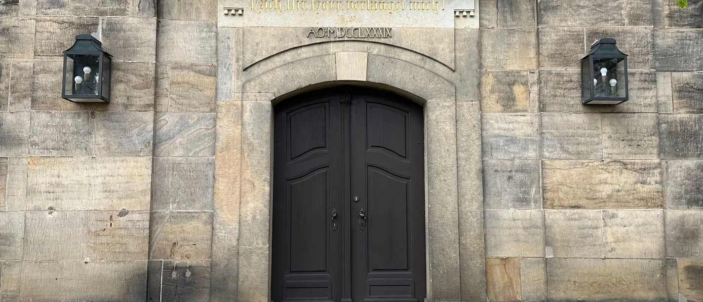 Tür der Kirche in Lohmen Dunkle Holztür in einem steinernen Gebäude, flankiert von zwei Laternen; darüber eine Inschrift in goldener Schrift.
