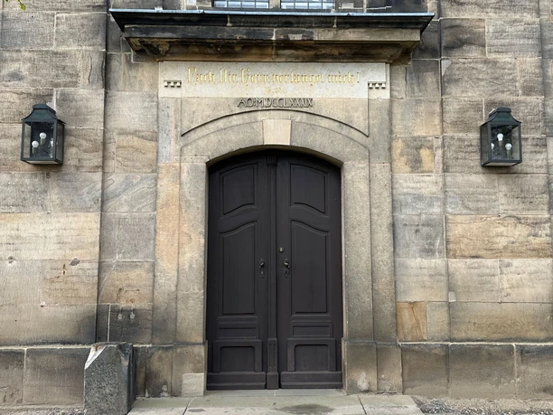 Tür der Kirche in Lohmen Dark wooden door in a stone building, flanked by two lanterns; above it an inscription in gold lettering.