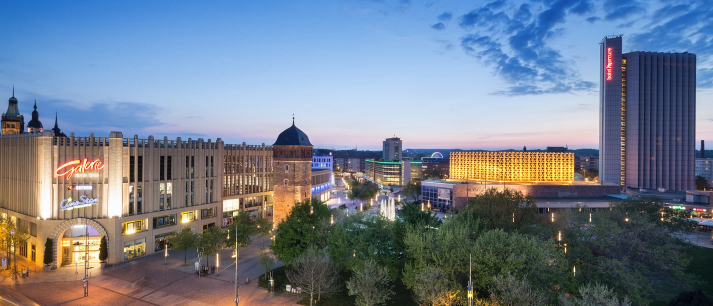 Chemnitz_City_Stadthallenpark_4094 (c) Dirk Hanus_CWE.jpg Stadthallenpark in Chemnitz bei Abenddämmerung, umgeben von beleuchteten Gebäuden und üppigen Grünflächen.