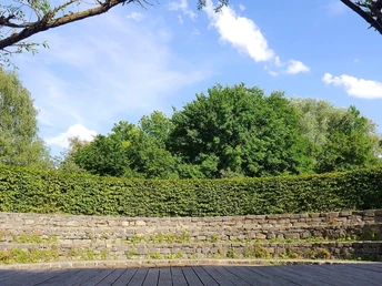 Steinernes Amphitheater im Grünen mit Holzpodest, umgeben von Bäumen und Hecken, unter blauem Himmel.