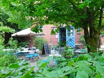 Mühlencafe am Umweltzentrum Heerser Mühle Grünes Café mit Außenbereich, Tischen und Sonnenschirm, umgeben von üppigem Pflanzenwuchs.