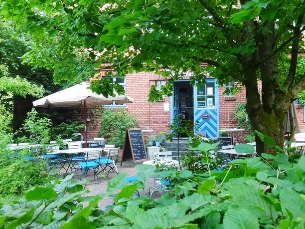 Mühlencafe am Umweltzentrum Heerser Mühle Grünes Café mit Außenbereich, Tischen und Sonnenschirm, umgeben von üppigem Pflanzenwuchs.