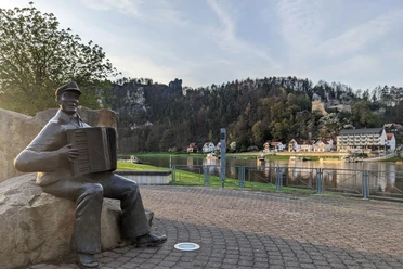 Musiker auf der Klangterrasse in Rathen Bronzestatue eines Mannes mit Akkordeon auf einem Platz, im Hintergrund Fluss, Häuser und bewaldeter Hügel mit Burgruine bei Sonnenuntergang.