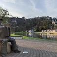 Musiker auf der Klangterrasse in Rathen Bronzestatue eines Mannes mit Akkordeon auf einem Platz, im Hintergrund Fluss, Häuser und bewaldeter Hügel mit Burgruine bei Sonnenuntergang.
