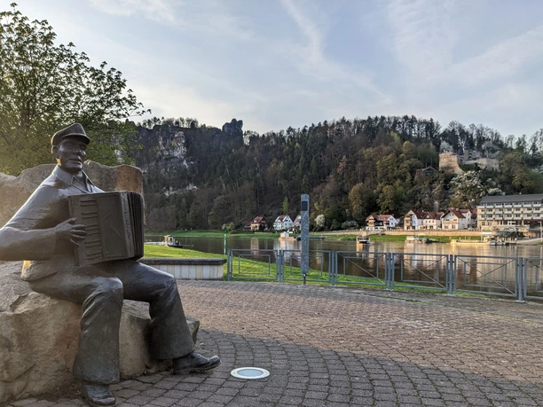 Musiker auf der Klangterrasse in Rathen Bronzestatue eines Mannes mit Akkordeon auf einem Platz, im Hintergrund Fluss, Häuser und bewaldeter Hügel mit Burgruine bei Sonnenuntergang.