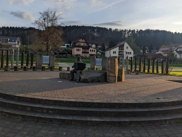 Stenen figuur van een accordeonspeler op een plein met omringende huizen en bomen op de achtergrond, in de schemering.