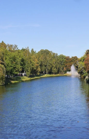 Radfahrer auf einem baumbestandenen Weg an einem breiten Wassergraben, umgeben von Grünflächen.