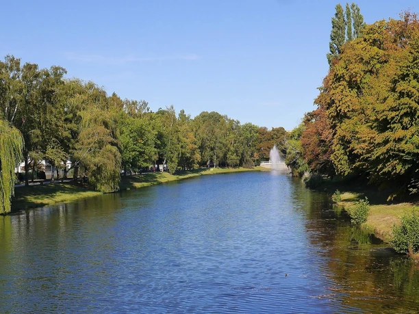An Wallanlagen in Herford entlang fahren Radfahrer auf einem baumbestandenen Weg an einem breiten Wassergraben, umgeben von Grünflächen.
