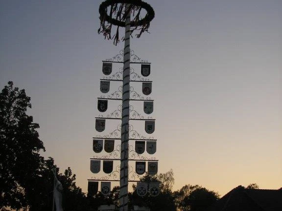 Ein Maibaum mit vielen Wappen steht vor einem Dorf bei Sonnenuntergang, umgeben von Bäumen.