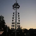 P4290808.jpg Ein Maibaum mit vielen Wappen steht vor einem Dorf bei Sonnenuntergang, umgeben von Bäumen.