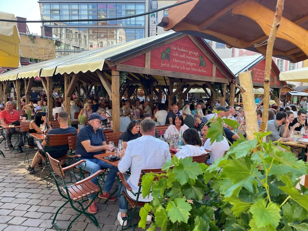 Weinfest Chemnitz 2.jpg Menschen genießen Wein und Geselligkeit unter schattigen Holzdächern auf einem Stadtfest.