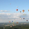 Heißluftballons in bunten Farben schweben über einer bewaldeten Landschaft unter blauem Himmel.