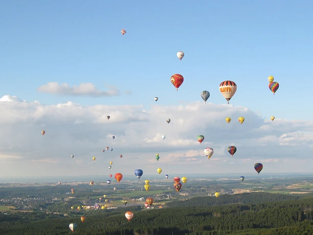 Heißluftballons in bunten Farben schweben über einer bewaldeten Landschaft unter blauem Himmel.