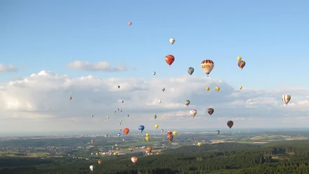 Rehm-Ballooning-1242x700.jpg Heißluftballons in bunten Farben schweben über einer bewaldeten Landschaft unter blauem Himmel.