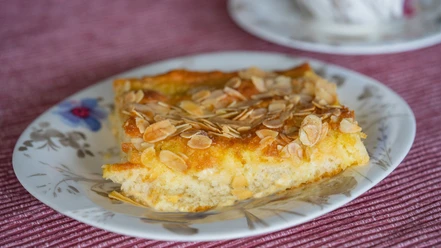 Butterkuchen-Ostfriesentee Ein Stück Butterkuchen mit Mandelsplittern liegt auf einem blumigen Teller vor einem farbigen Tischtuch.