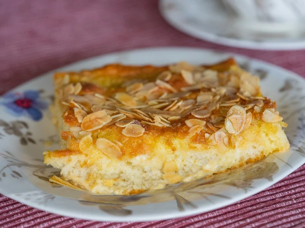 Butterkuchen-Ostfriesentee Ein Stück Butterkuchen mit Mandelsplittern liegt auf einem blumigen Teller vor einem farbigen Tischtuch.