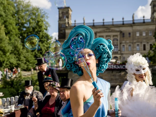 k-Dresdner Schlössernacht 2022 Foto Michael Schmidt Motiv 13.jpg Frau mit blauer Perücke pustet Seifenblasen bei einem Kostümfest vor einem Schloss im Freien.