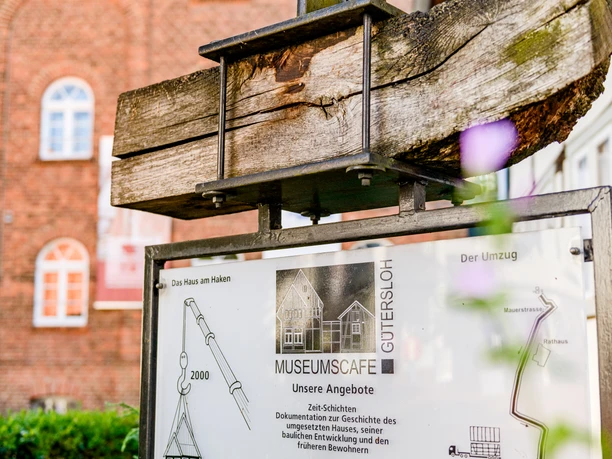 Holzstab mit Metallschild vor Backsteingebäude; Infotafel zu Museumscafé in Gütersloh.