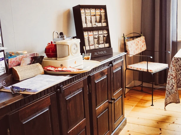 cafe-harms-soderstorf1.jpg Holzschrank mit Teeauswahl, Retroradio und Speisekarte neben einem Tisch mit floraler Decke.