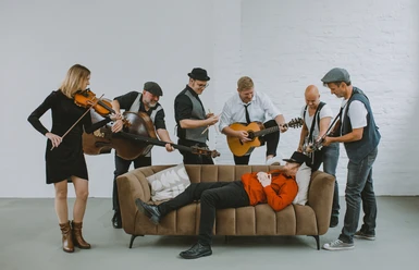Eine sitzende Band spielt fröhlich um einen auf dem Sofa liegenden Mann, alle in legerer Kleidung.