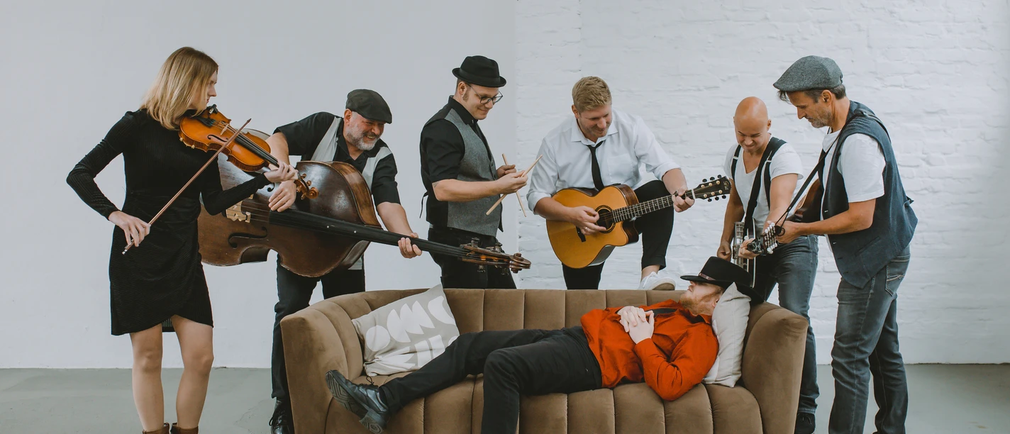 Eine sitzende Band spielt fröhlich um einen auf dem Sofa liegenden Mann, alle in legerer Kleidung.