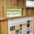 Holzhütte mit Informationstafel über Vogelarten. Blick durch Fenster auf Weiher und grüne Natur.
