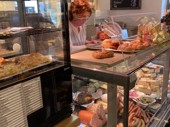 Dorfladen Riegsee Verkäuferin im Dorfladen schneidet Aufschnitt neben Gebäck und Menütafel.