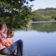 Senne-See©Stadt SHS (1).jpg Mann und Frau sitzen am Seeufer, umgeben von Bäumen, mit Blick auf das ruhige Wasser des Sees.