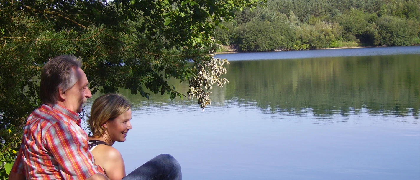 Senne-See©Stadt SHS (1).jpg Mann und Frau sitzen am Seeufer, umgeben von Bäumen, mit Blick auf das ruhige Wasser des Sees.