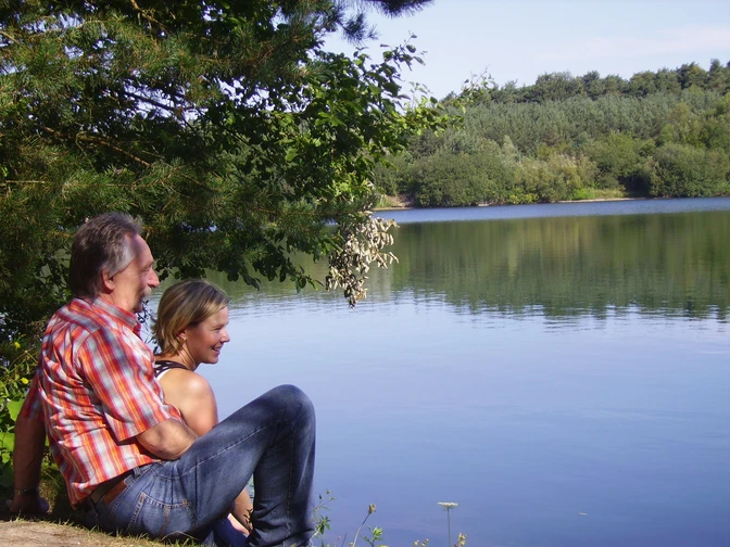 Senne-See©Stadt SHS (1).jpg Mann und Frau sitzen am Seeufer, umgeben von Bäumen, mit Blick auf das ruhige Wasser des Sees.