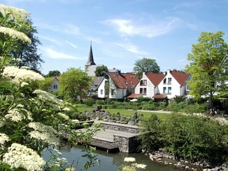 Blick über den Angergarten in Wülfrath Der Angergarten in Wülfrath mit historischen Häusern und einem Steg am Wasser, umgeben von Grün.