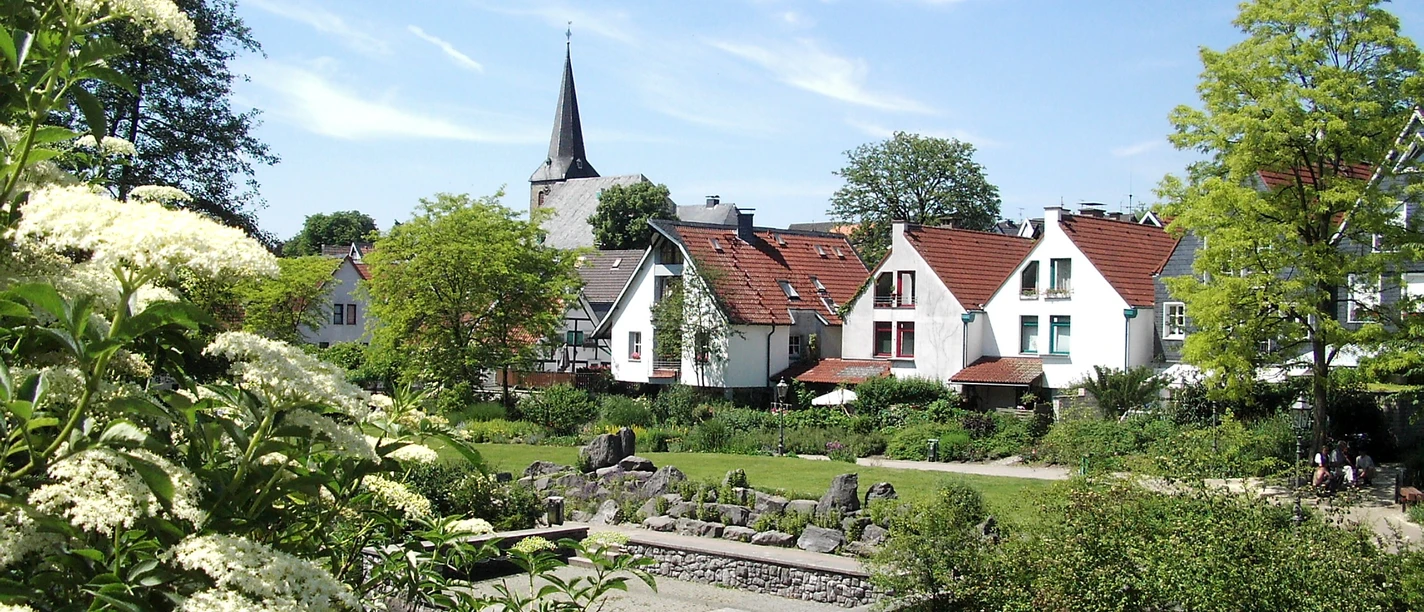 Blick über den Angergarten in Wülfrath Der Angergarten in Wülfrath mit historischen Häusern und einem Steg am Wasser, umgeben von Grün.
