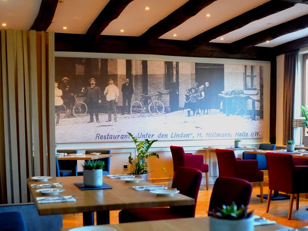 Modernes Restaurant mit Holzbalkendecke, bunten Stühlen und Wandbild historischer Straßenszene.