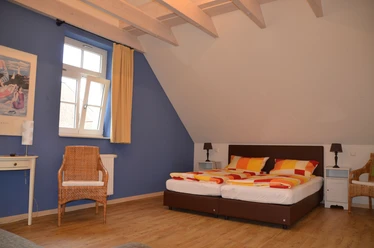 Ein gemütliches Doppelzimmer mit blauen Wänden, Holzdecke, einem Fenster und orange-gelben Bettdecken.
